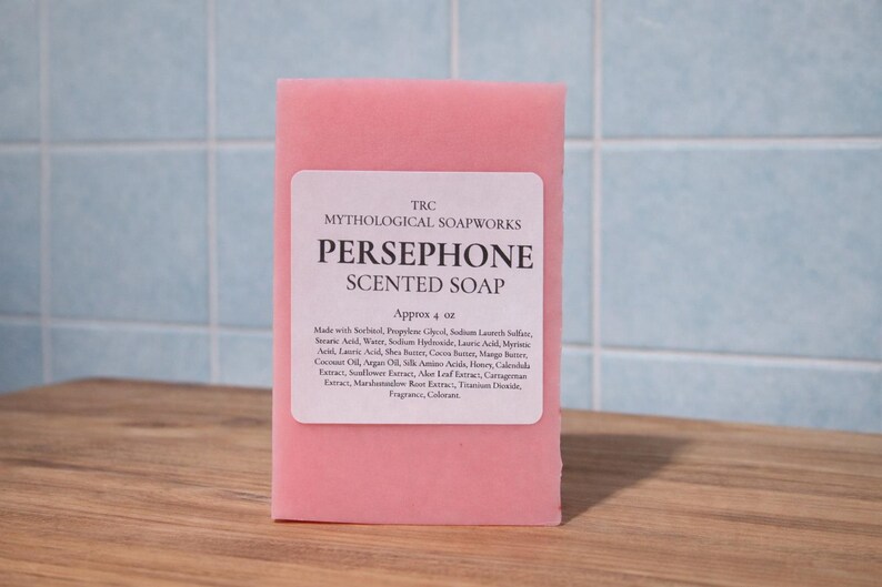 May include: A pink bar of Persephone scented soap from Mythological Soapworks. The soap has a white label with the product name and ingredients. The bar is approximately 4 oz. and sits on a wooden surface against a blue tiled background.