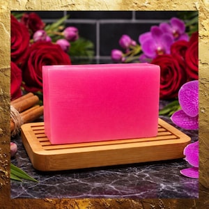 May include: A vibrant pink soap bar rests on a natural bamboo soap dish. The soap is surrounded by red roses, purple orchids, and cinnamon sticks, creating a spa-like aesthetic. The soap dish has a rectangular shape with parallel grooves.