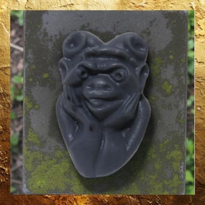 May include: A dark gray gargoyle sculpture with a detailed face, hands supporting the chin, and curled horns. The sculpture is mounted on a weathered, moss-covered stone surface, set against a gold-toned background.