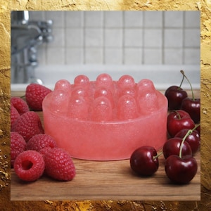 May include: A round, pink soap bar with a textured surface, surrounded by fresh raspberries and cherries on a wooden surface. The soap has a bubbly appearance and is a vibrant shade of pink.