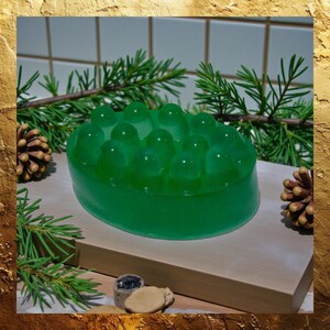 May include: A green oval-shaped soap with a textured top, resting on a wooden surface. The soap is surrounded by pine needles and pine cones, suggesting a natural, botanical theme. The soap's design features rounded protrusions.