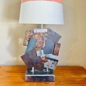 May include: A modern table lamp with a two-toned lampshade. The shade is coral pink on top and white on the bottom. The base is made of metal and has a geometric design with a rusty finish.