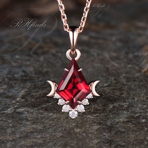 May include: A rose gold necklace with a pendant featuring a red gemstone surrounded by smaller clear gemstones. The pendant is shaped like a triangle with crescent moons on either side.