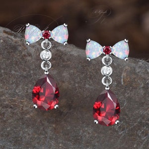 Può includere: Un paio di orecchini d'argento con una pietra rossa a forma di lacrima e un fiocco di opale bianco. Gli orecchini sono pendenti e hanno un piccolo accento di pietra chiara.