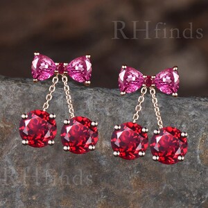 May include: A pair of cherry-shaped earrings. Each earring features a pink bow at the top, connected by a delicate chain to two round, deep red gemstones. The earrings are set against a neutral background.