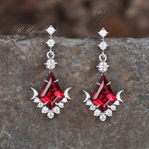 May include: A pair of silver dangle earrings with a red gemstone and clear cubic zirconia stones. The earrings feature a crescent moon design.