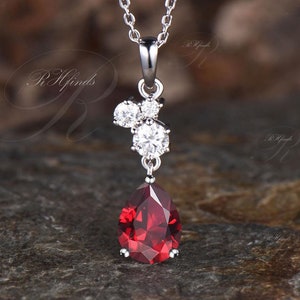 May include: A silver chain necklace with a teardrop-shaped red gemstone pendant. The pendant is surrounded by smaller clear gemstones.