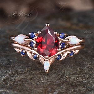 May include: A pear-shaped red gemstone engagement ring set with a delicate band featuring white opals and blue sapphires.