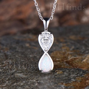 Puede incluir: Un collar de tono plateado con un colgante de dos piedras en forma de pera dispuestas en un bucle vertical similar al infinito. La piedra superior es una gema transparente facetada, mientras que la inferior es una ópalo blanco lechoso.