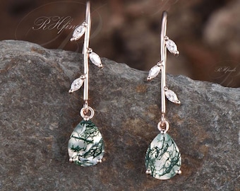 May include: A pair of rose gold earrings featuring a delicate branch design and green moss agate teardrop stones. The earrings are accented with small clear cubic zirconia stones.
