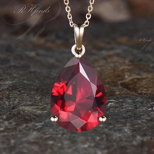 May include: A gold-toned necklace featuring a faceted, teardrop-shaped, deep red pendant. The pendant is suspended from a delicate chain and has a gold-colored setting. The gemstone is a vibrant, eye-catching red.