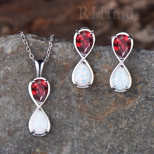 May include: A jewelry set consisting of a pendant necklace and matching earrings. Each piece features a silver-toned infinity-style frame holding a pear-shaped red garnet gemstone above a pear-shaped white opal gemstone. The text RHfinds is visible.