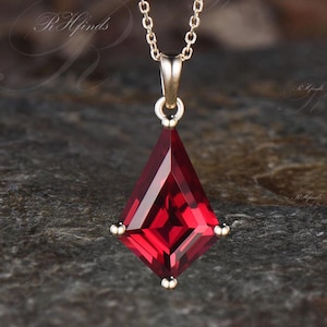 May include: A gold-colored chain necklace with a red, diamond-shaped pendant. The pendant has a faceted surface and small gold accents at each corner. The necklace is displayed on a dark, textured surface.