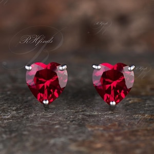 以下が含まれることがあります： 赤いキュービックジルコニアの宝石が付いた、銀製のハート型のスタッドピアスのペアです。