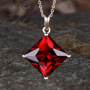May include: A gold-colored necklace with a square, deep red gemstone pendant. The pendant is a faceted, princess-cut stone with gold accents at each corner. The chain is delicate and hangs from a matching gold bail.