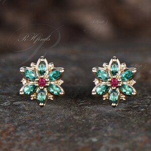 May include: A pair of gold earrings with a flower design. Each earring features a red center stone surrounded by green stones.