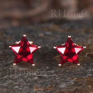 May include: A pair of star-shaped stud earrings. Each earring features a vibrant red gemstone set in a gold-colored metal. The earrings are displayed on a dark, textured surface.