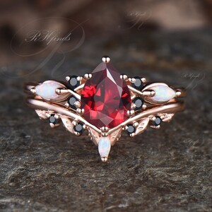 May include: A pear-shaped red gemstone ring set in rose gold with black and white gemstones on the band. The ring is on a gray background.