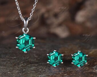 May include: A silver necklace with a hexagonal emerald pendant and matching stud earrings. The jewelry set features faceted, vibrant green gemstones. The necklace chain and earring posts are silver. The set is displayed on a textured, dark surface.