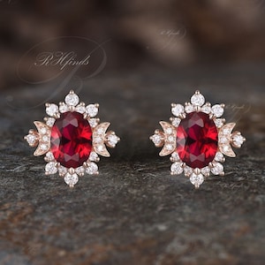 Puede incluir: Un par de pendientes con una piedra ovalada roja rodeada por un halo de piedras blancas más pequeñas. Los pendientes tienen un diseño floral delicado.