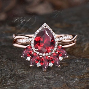 Puede incluir: Un anillo de piedra roja en forma de pera con un halo de diamantes más pequeños. El anillo está engastado en oro rosa y tiene una banda a juego con un diseño delicado.