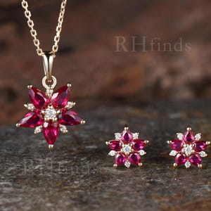 May include: A gold necklace and matching stud earrings set featuring flower-shaped pendants. Each piece contains pear-cut deep red gemstones arranged as petals around a central round white stone. The text RHfinds is visible in the background.