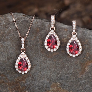 May include: A set of rose gold jewellery featuring a teardrop-shaped pendant and earrings with a red gemstone surrounded by clear cubic zirconia stones.