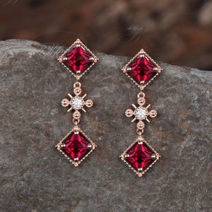以下が含まれることがあります： 赤い正方形の宝石と、十字架のデザインの中心に小さなダイヤモンド型の宝石が付いた、ぶら下がりのイヤリングのペア。イヤリングはローズゴールド製です。