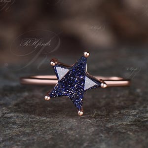 May include: A rose gold ring featuring a star-shaped blue gemstone with sparkling inclusions. The star has five points, with small gold accents at each point. The band is a thin, polished rose gold.