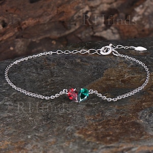 May include: A delicate silver bracelet featuring two heart-shaped gemstones, one red and one green. The bracelet has a fine chain and a secure clasp. The gemstones are set close together, creating a charming and elegant design.