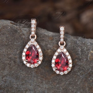 May include: A pair of rose gold dangle earrings with a pear-shaped red gemstone surrounded by a halo of smaller clear gemstones.