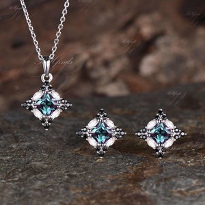 May include: A silver necklace and matching earrings set. Each piece features a square design with a central teal stone, surrounded by white and black accents. The necklace has a delicate chain.