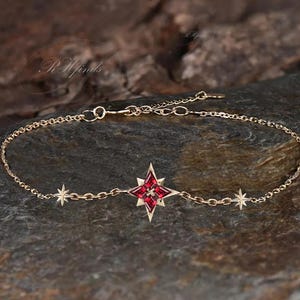 May include: Gold-toned bracelet featuring a central red starburst design and smaller star accents. The delicate chain is displayed on a dark, textured surface, highlighting the jewelry's intricate details. The bracelet is a piece of fine jewelry.