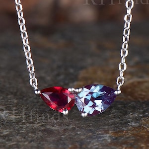 May include: A silver-toned chain necklace featuring two pear-cut gemstones, one deep red and one multifaceted blue-purple, set side-by-side. The text RHfinds is visible in the background. The delicate metal chain connects to each side of the stones.