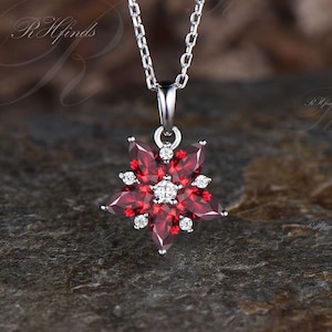 May include: A silver-toned necklace featuring a flower-shaped pendant. The pendant is composed of five marquise-cut deep red stones arranged as petals, accented with small clear round stones between the petals and a single clear stone at the center.