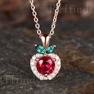 May include: A rose gold necklace with a pendant shaped like an apple. The apple pendant features a large, round, red gemstone surrounded by smaller white gemstones. Two green leaf-shaped gemstones sit above the red gemstone.