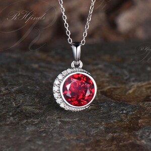 May include: A silver chain necklace with a pendant featuring a large, round, red gemstone surrounded by smaller, clear gemstones.