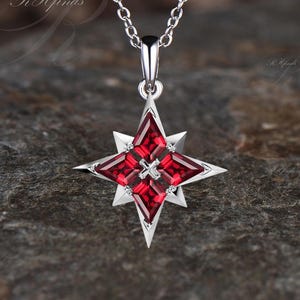 May include: A silver-toned pendant necklace featuring a starburst design. The starburst has a central square and four red gemstone sections, with small white accents. The necklace chain is delicate and silver, suitable for everyday wear.