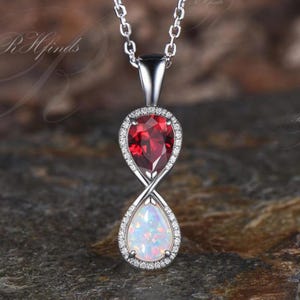 May include: A silver-toned pendant necklace featuring an infinity-shaped design. The top section holds a pear-cut deep red gemstone, while the bottom section contains a pear-cut iridescent white opal. Both stones are framed by small clear stones.