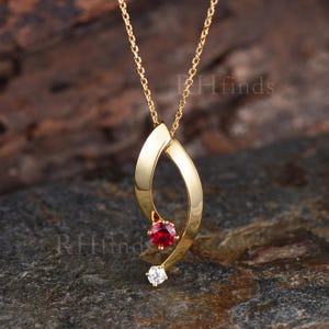 May include: A gold-toned necklace featuring a curved, polished metal pendant suspended from a delicate chain. The pendant is accented with a single round, faceted red gemstone and a smaller, clear round gemstone at the bottom tip. Text reads RHfinds.
