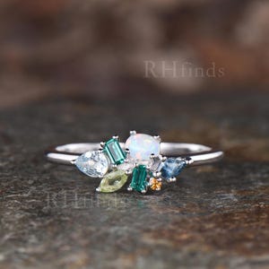 May include: A silver-toned ring featuring a cluster of multi-colored gemstones including a central oval white opal, two rectangular green stones, a pear-shaped light blue stone, a marquise-cut light green stone, and small blue and orange accents.