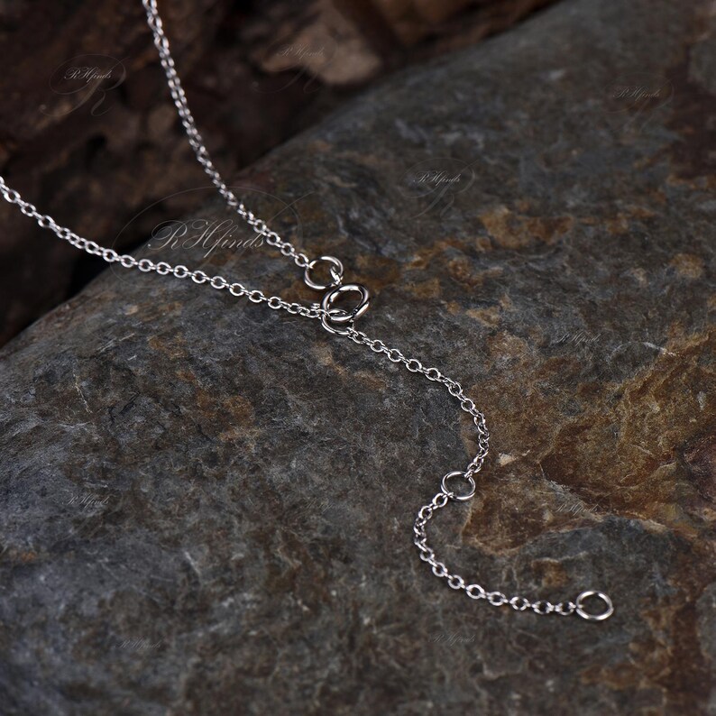 May include: Close-up of a delicate silver necklace chain with a small clasp and adjustable length. The chain is displayed on a dark, textured stone surface, highlighting its intricate design and reflective qualities.