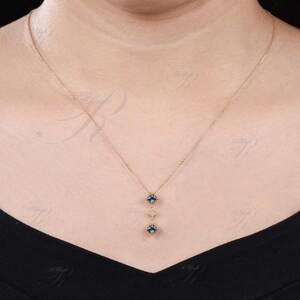 Op de afbeelding: Een delicate gouden ketting met een hanger met drie blauwe edelstenen en een kleinere witte edelsteen. De hanger heeft de vorm van een verticale lijn met de edelstenen symmetrisch gerangschikt.