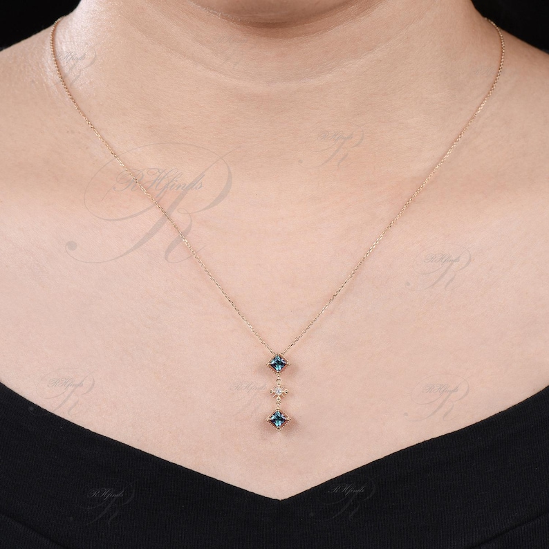 Op de afbeelding: Een delicate gouden ketting met een hanger met drie blauwe edelstenen en een kleinere witte edelsteen. De hanger heeft de vorm van een verticale lijn met de edelstenen symmetrisch gerangschikt.