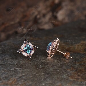 May include: Rose gold stud earrings with a square, teal-colored center stone, surrounded by black and white accent stones. The earrings have a floral design and are set on a dark, textured surface.