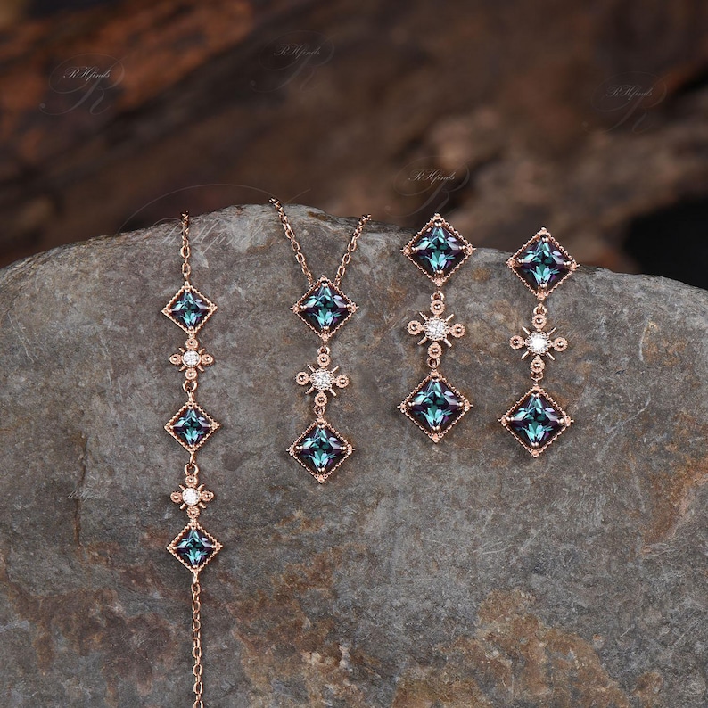 Op de afbeelding: Een set van drie gouden sieraden met blauwe en heldere edelstenen. De set bevat een ketting, een armband en een paar oorbellen. Elk stuk heeft een vierkante hanger met een blauwe edelsteen omringd door kleinere heldere edelstenen.