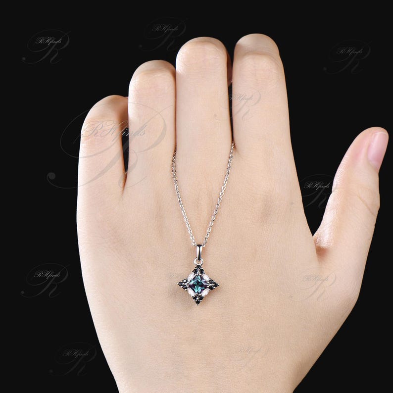 May include: A silver necklace with a square pendant featuring a central teal gemstone surrounded by smaller black and white stones. The pendant hangs from a delicate chain, displayed against a neutral background.