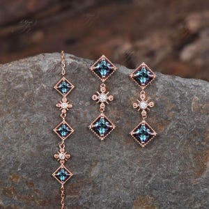 Op de afbeelding: Een set van drie gouden oorbellen met blauwe en heldere edelstenen. De oorbellen hebben een vierkante edelsteen met een kleinere, heldere edelsteen aan elke kant. De oorbellen zijn verbonden door een ketting met een kleine, heldere edelsteen in het midden.
