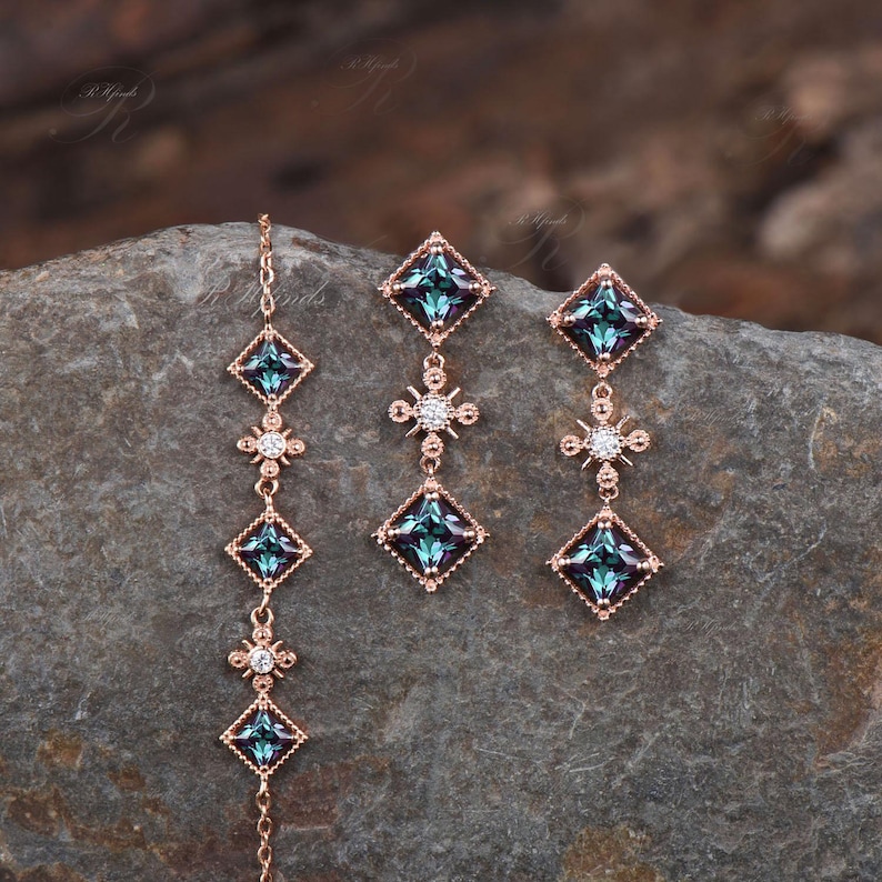 Op de afbeelding: Een set van drie gouden oorbellen met blauwe en heldere edelstenen. De oorbellen hebben een vierkante edelsteen met een kleinere, heldere edelsteen aan elke kant. De oorbellen zijn verbonden door een ketting met een kleine, heldere edelsteen in het midden.