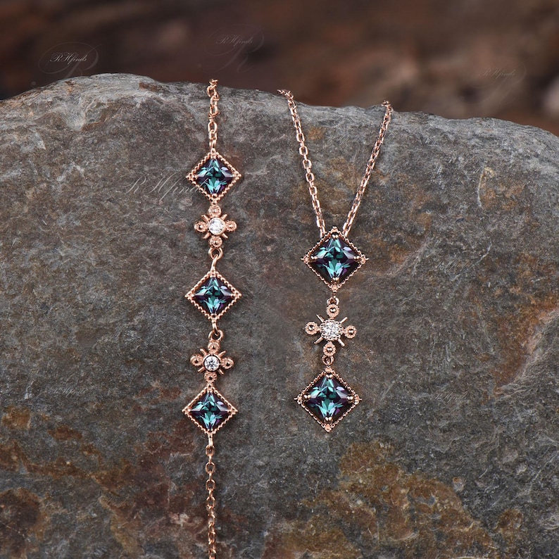 Op de afbeelding: Twee delicate kettingen met een ros&eacute;gouden ketting en blauwe edelsteen hangers. De hangers zijn vierkant met een kleiner diamantvormig accent in het midden. De kettingen liggen op een grijs, getextureerd oppervlak.